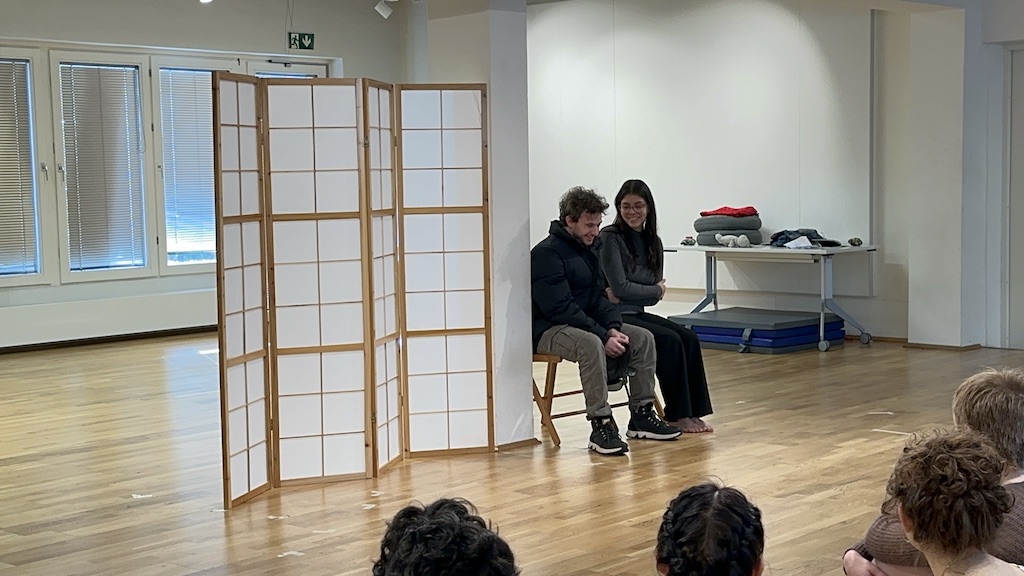 Two actors sit next to each other smiling in mid-performance of 'Faith Hope Charity'. A few students sit in the audience watching the performance.